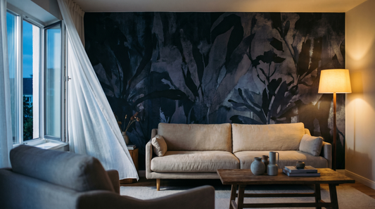A wide editorial interior photograph (16:9) of a summer living room at dusk: a bold abstract botanical accent wall in deep